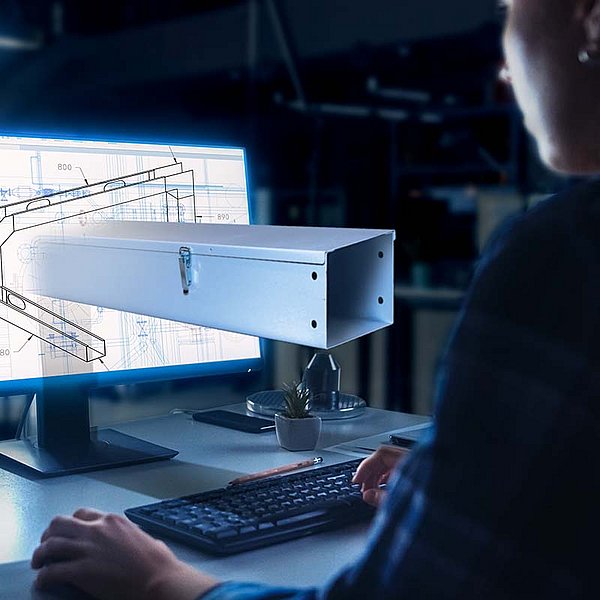 A woman draws a component assembly on the computer, while a section of cable trunking protrudes from the screen.