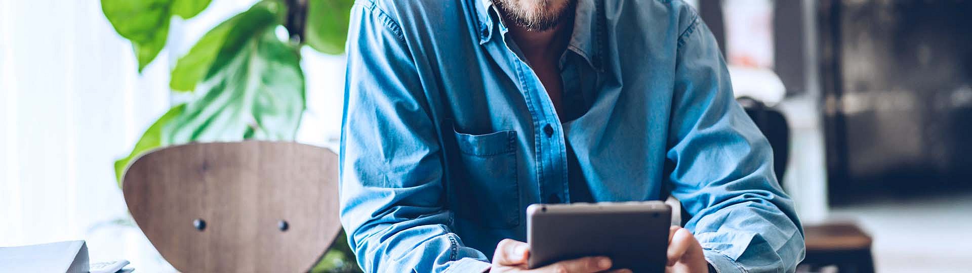 Ein Mann im blauen Hemd sitzt auf einem Stuhl und schaut auf sein Tablet